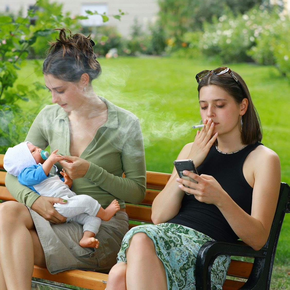 Zwei Frauen sitzen im Garten. Eine stillt ein Baby, die andere raucht und schaut auf ihr Handy.